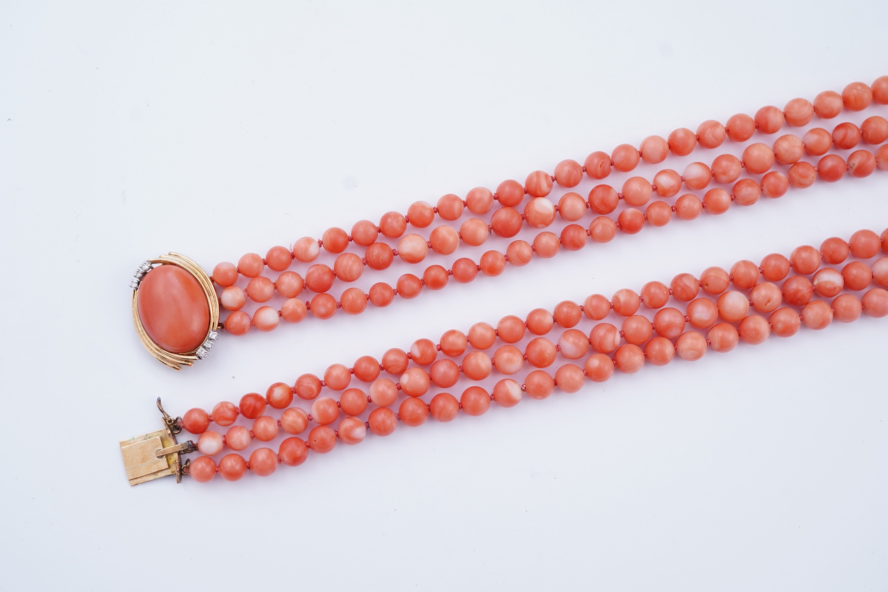 A coral necklace, composed of three strands of coral beads measuring approximately 5.1-6.6mm diameter, to a gold clasp set with a coral cabochon and brilliant-cut diamonds, mounted in gold, length 45.5cm, stamped 14K, ca